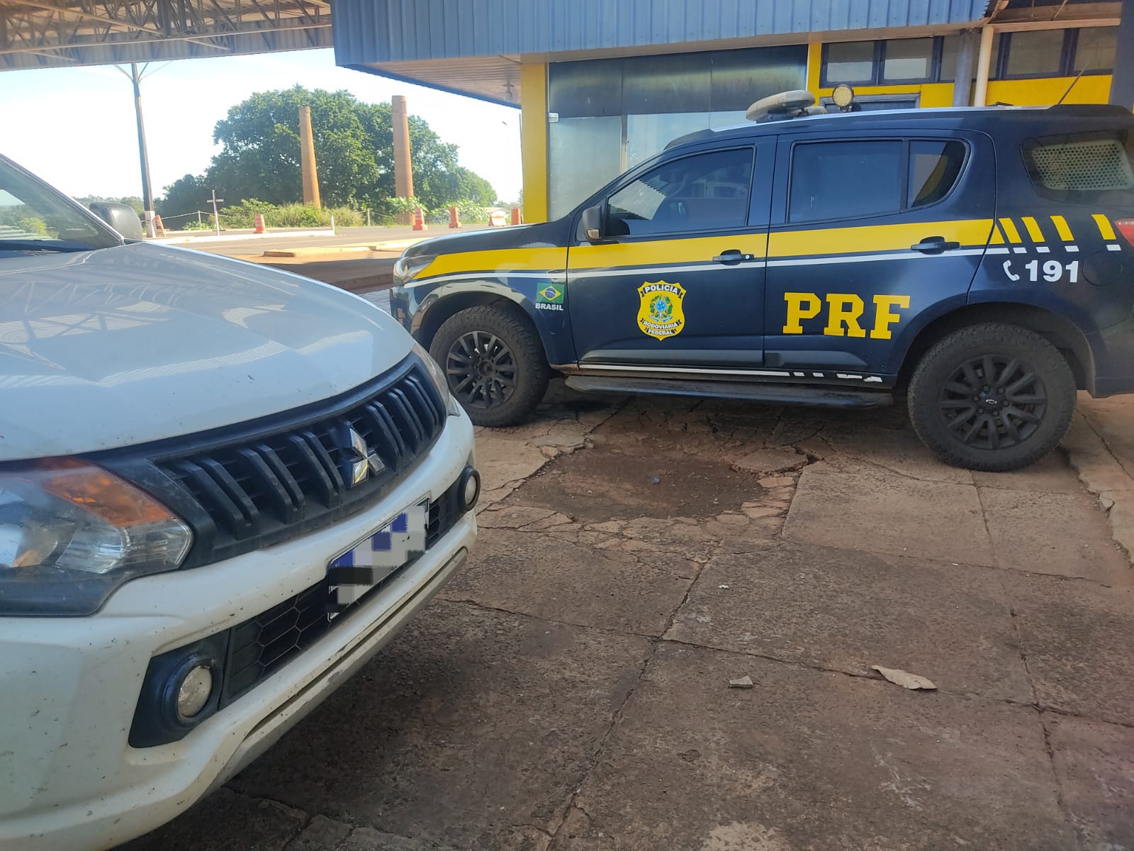 Uma imagem em plano médio e ângulo normal, capturada em uma área externa pavimentada e parcialmente coberta, mostra a lateral de uma viatura policial e a parte frontal de uma caminhonete branca.