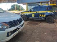 Uma imagem em plano médio e ângulo normal, capturada em uma área externa pavimentada e parcialmente coberta, mostra a lateral de uma viatura policial e a parte frontal de uma caminhonete branca.