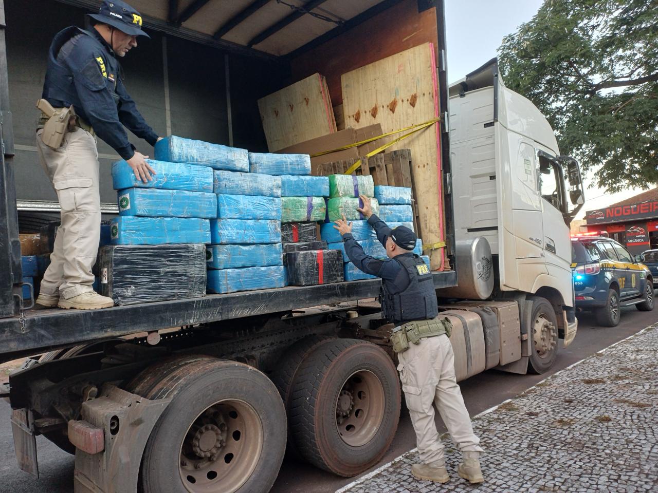 Dois policiais rodoviários federais fiscalizam um caminhão baú estacionado às margens da rodovia. Na carroceria aberta, dezenas de fardos de maconha embalados em plástico azul, verde e preto estão empilhados. Um policial está sobre o caminhão manuseando os volumes, enquanto outro, no solo, auxilia na inspeção da carga. Ao fundo, uma viatura da PRF permanece estacionada próxima ao local da apreensão.