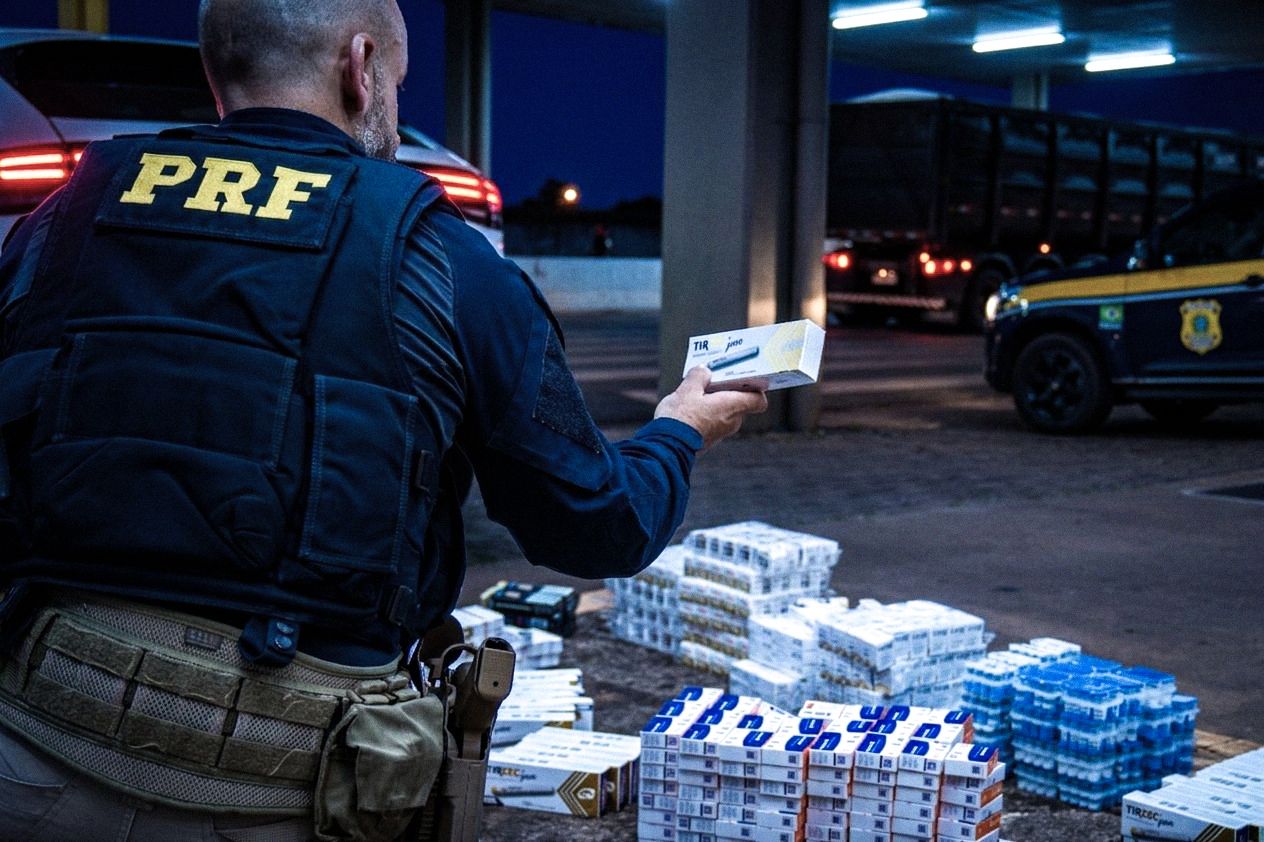Policial rodoviário federal de costas examina uma caixa de medicamento emagrecedor (Tirzepatida) durante uma operação noturna. No chão, à sua frente, há uma grande quantidade de caixas do mesmo medicamento empilhadas. Ao fundo, sob a iluminação de um posto de fiscalização, veem-se uma viatura da PRF e um caminhão.