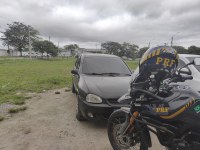A imagem mostra um carro preto e uma motocicleta da PRF na frente do veículo