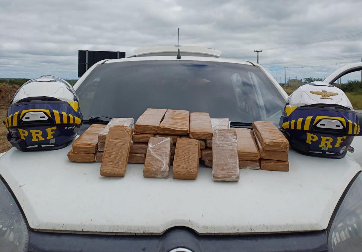 A imagem apresenta 29 tabletes de maconha em cima do capô de um carro branco. Ao lado da droga há dois capacetes