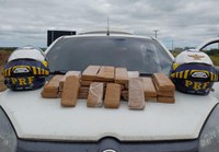 A imagem apresenta 29 tabletes de maconha em cima do capô de um carro branco. Ao lado da droga há dois capacetes