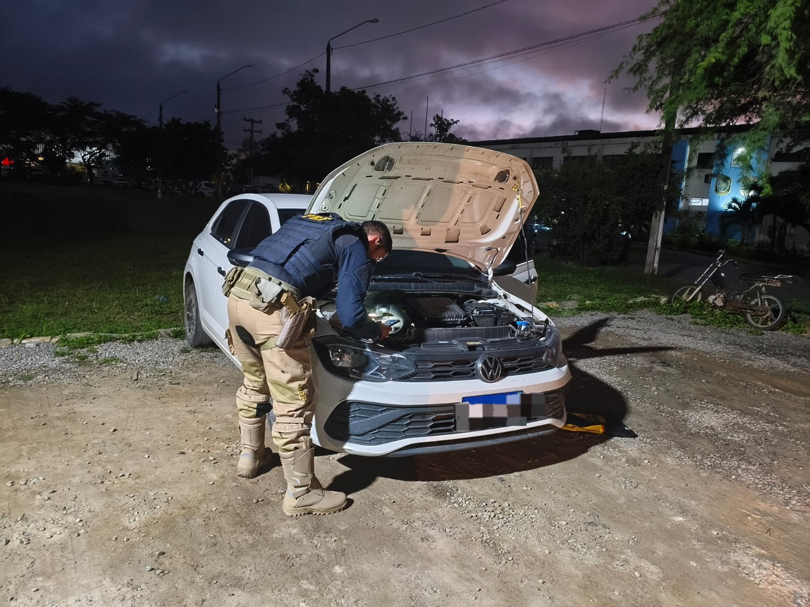 A imagem mostra um policial rodoviário federal verificando o motor de um carro