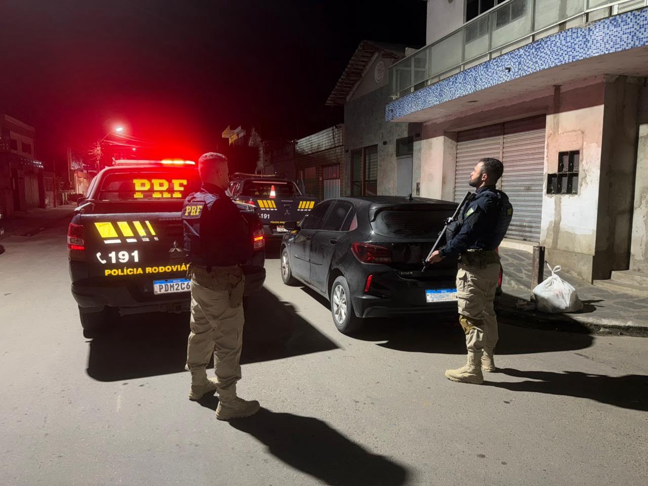 Foto noturna de uma rua urbana. Dois agentes da polícia, usando uniforme escuro e calças bege, estão posicionados ao lado de um carro preto hatchback parado na via. Um dos agentes está à esquerda, próximo a uma caminhonete da polícia com luzes vermelhas de emergência acesas; o outro está à direita, observando a cena e segurando uma arma longa apontada para baixo. Ao fundo, há outra viatura policial estacionada. As luzes vermelhas iluminam a rua e refletem nos veículos. À direita da imagem aparecem fachadas de prédios com portas metálicas fechadas e uma sacola de lixo sobre a calçada. O ambiente é escuro, com iluminação vinda dos postes e das viaturas.