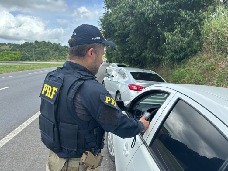 Um agente da Polícia Rodoviária Federal (PRF), vestindo uniforme escuro e colete com as letras amarelas “PRF”, está parado no acostamento de uma rodovia. Ele segura um aparelho portátil próximo à janela aberta de um carro prata. Atrás desse veículo, há outro carro estacionado no acostamento. Ao lado da estrada há vegetação densa e árvores, enquanto o céu está parcialmente nublado com nuvens escuras.