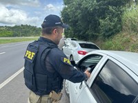 Um agente da Polícia Rodoviária Federal (PRF), vestindo uniforme escuro e colete com as letras amarelas “PRF”, está parado no acostamento de uma rodovia. Ele segura um aparelho portátil próximo à janela aberta de um carro prata. Atrás desse veículo, há outro carro estacionado no acostamento. Ao lado da estrada há vegetação densa e árvores, enquanto o céu está parcialmente nublado com nuvens escuras.