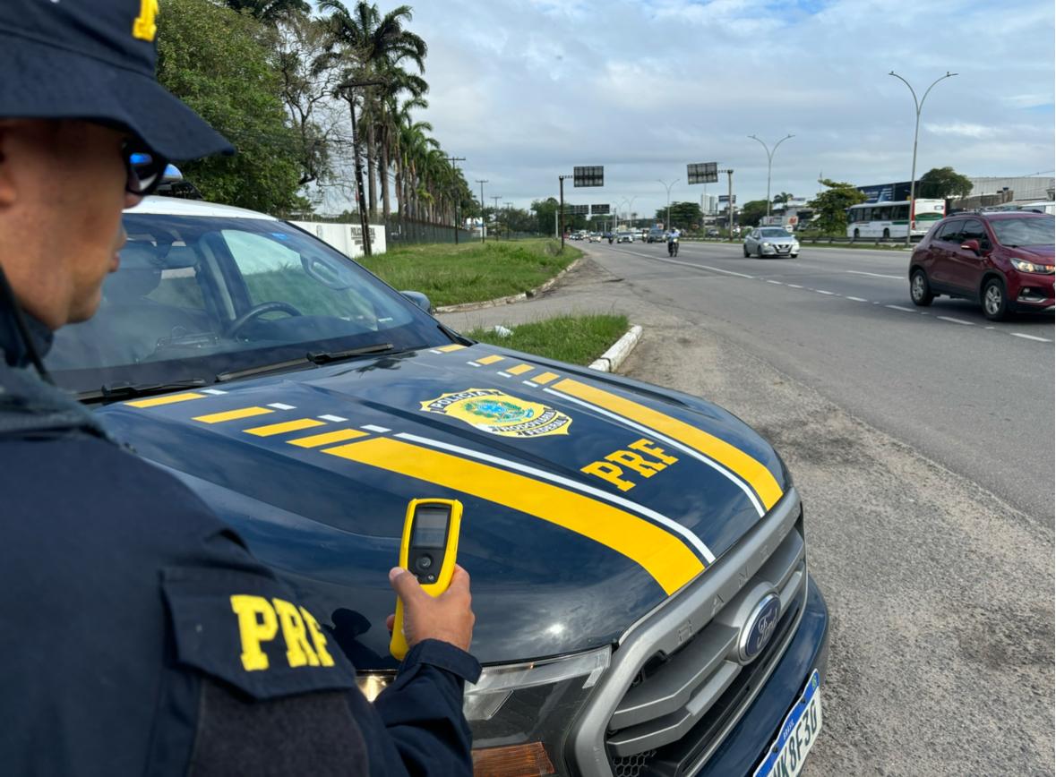 Fotografia de uma operação da Polícia Rodoviária Federal (PRF) realizada às margens de uma rodovia de múltiplas faixas. Em primeiro plano, um policial rodoviário federal aparece parcialmente de perfil, segurando um equipamento portátil de fiscalização. Ao lado dele está uma viatura da PRF, com a identificação institucional e o brasão da corporação destacados no capô. Ao fundo, observa-se o fluxo de veículos na via, incluindo carros, motocicletas e ônibus, além de canteiro central gramado, palmeiras e placas de sinalização. O céu está nublado, e a cena retrata uma atividade de monitoramento e fiscalização voltada à segurança viária.