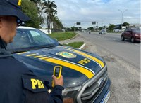 Fotografia de uma operação da Polícia Rodoviária Federal (PRF) realizada às margens de uma rodovia de múltiplas faixas. Em primeiro plano, um policial rodoviário federal aparece parcialmente de perfil, segurando um equipamento portátil de fiscalização. Ao lado dele está uma viatura da PRF, com a identificação institucional e o brasão da corporação destacados no capô. Ao fundo, observa-se o fluxo de veículos na via, incluindo carros, motocicletas e ônibus, além de canteiro central gramado, palmeiras e placas de sinalização. O céu está nublado, e a cena retrata uma atividade de monitoramento e fiscalização voltada à segurança viária.