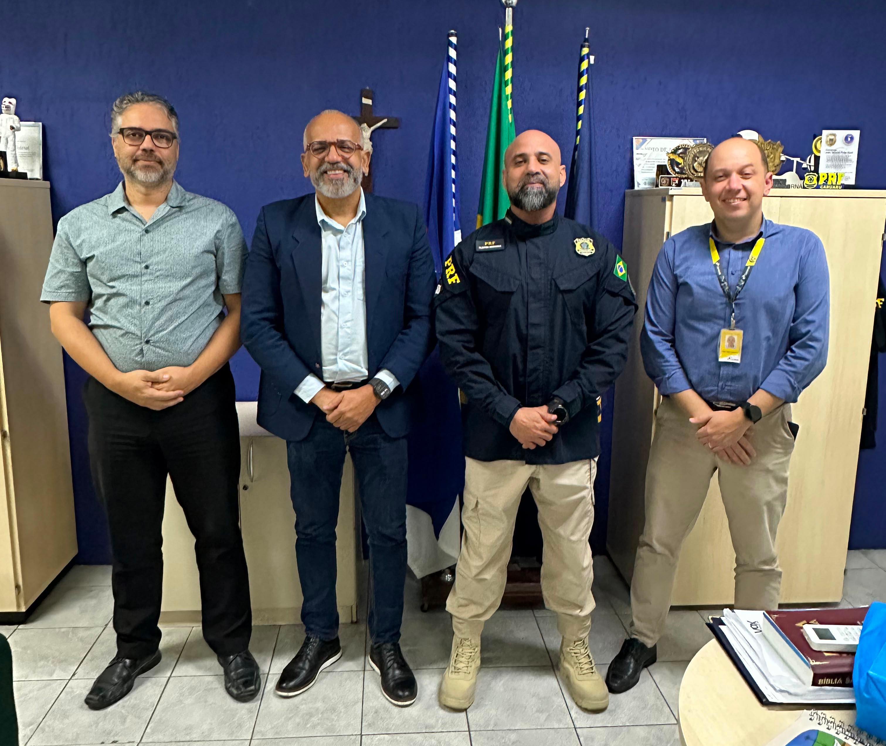 Quatro homens posam lado a lado em um escritório, diante de bandeiras e de uma cruz na parede. Um deles veste uniforme da Polícia Rodoviária Federal (PRF), enquanto os demais usam roupas sociais ou esporte fino. Todos estão de frente para a câmera em um registro institucional.