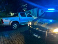Foto noturna de uma viatura da Polícia Rodoviária Federal (PRF), com as luzes de emergência azuis acesas, estacionada ao lado de uma caminhonete prata. Os veículos estão em frente a um imóvel, em um registro de ação policial.
