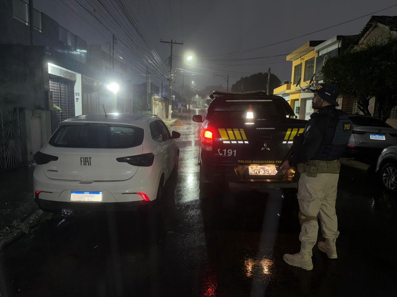 Foto noturna de uma rua molhada pela chuva. À esquerda, um carro branco está estacionado ao lado de uma caminhonete da Polícia Rodoviária Federal. À direita da imagem, um agente da PRF, vestindo uniforme escuro, colete tático e calças bege, permanece de pé observando a via. A viatura policial exibe luzes traseiras acesas e sinalização refletiva amarela. O asfalto úmido reflete as luzes dos postes e dos veículos. Ao fundo, há casas e prédios residenciais, postes com fiação aérea e uma atmosfera de neblina ou garoa, que reduz a visibilidade e reforça o clima noturno.
