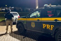Foto noturna de uma abordagem da Polícia Rodoviária Federal (PRF). Em primeiro plano, à direita, há uma viatura da PRF nas cores preta e amarela, com a sigla "PRF" em destaque na lateral e o giroflex no teto. À esquerda, um policial está de costas, usando uniforme tático, enquanto conversa ou realiza uma fiscalização em um carro branco parado à frente. Ao fundo, há iluminação de prédios e veículos, com a cena ocorrendo em uma via pavimentada.