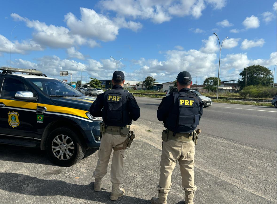 Dois policiais rodoviários federais ao lado da viatura de frente a rodovia.