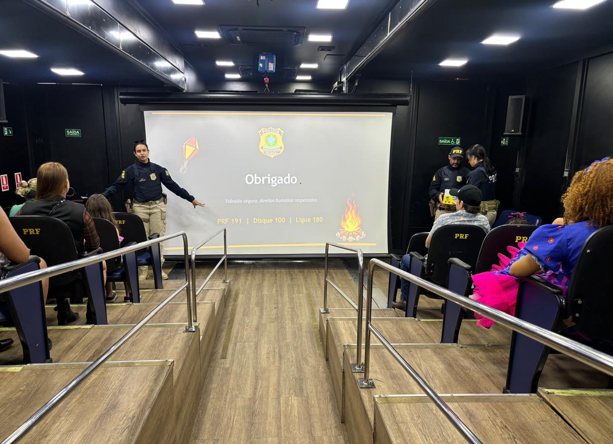 Policial da PRF conduz atividade educativa em auditório móvel, diante de público sentado.