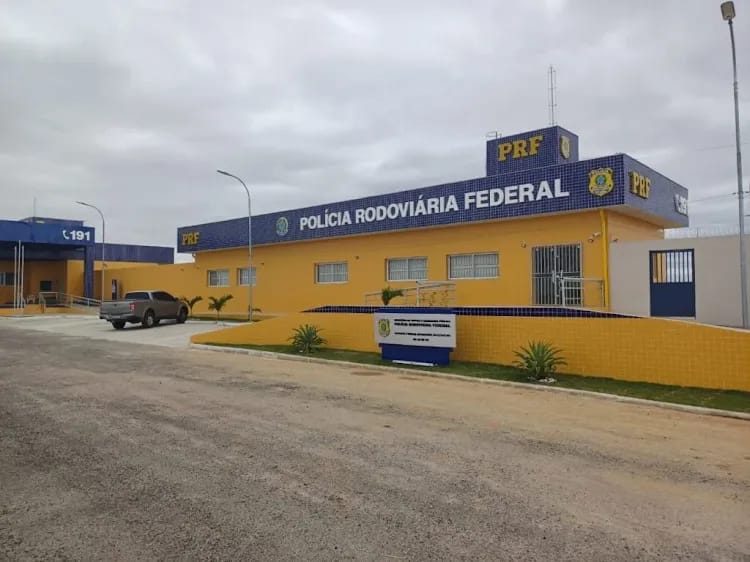 Fotografia diurna da sede da Polícia Rodoviária Federal (PRF), em um prédio térreo nas cores amarelo e azul. Na fachada, destaca-se a inscrição “Polícia Rodoviária Federal”, acompanhada do brasão e da sigla PRF. À esquerda, há uma estrutura identificada com o número de emergência 191 e uma caminhonete estacionada. Na parte frontal, um muro baixo amarelo, com detalhes azuis, delimita a área da unidade, onde também há pequenas plantas ornamentais. O céu está nublado, e a rua em frente ao prédio é pavimentada com paralelepípedos.