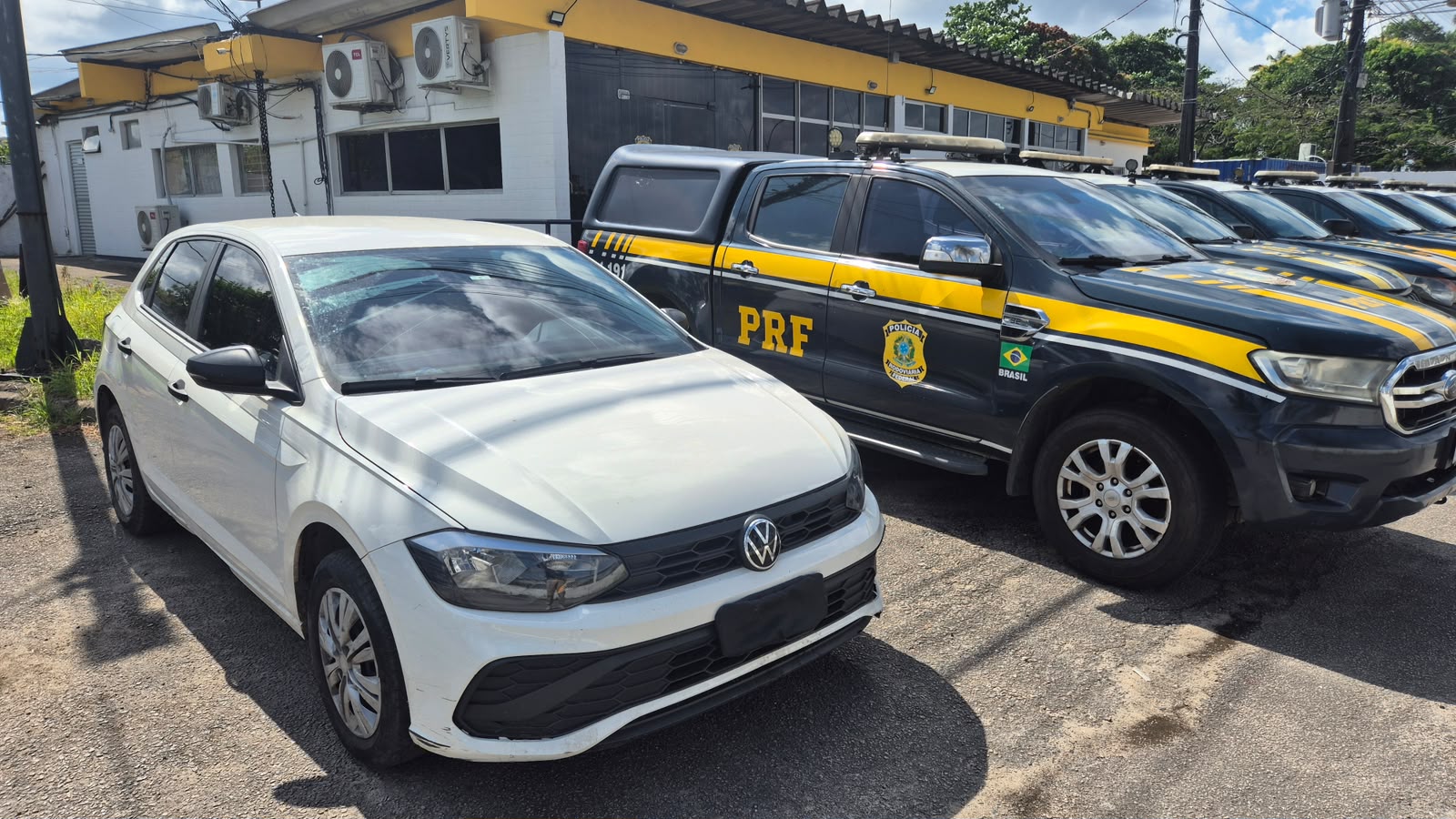 Fotografia diurna de um pátio da Polícia Rodoviária Federal (PRF). Em primeiro plano, há um carro branco da Volkswagen, visto de frente e pela lateral, com a placa encoberta. Ao lado e ao fundo, várias caminhonetes oficiais da PRF estão estacionadas em fila, na cor azul-escura, com faixas amarelas, brasões e identificação da instituição. Os veículos estão em frente a um prédio de fachada branca e amarela, com janelas amplas, aparelhos de ar-condicionado e cobertura escura. A luz do sol ilumina o local e projeta sombras dos veículos sobre o chão. Há árvores e vegetação nas laterais do terreno.