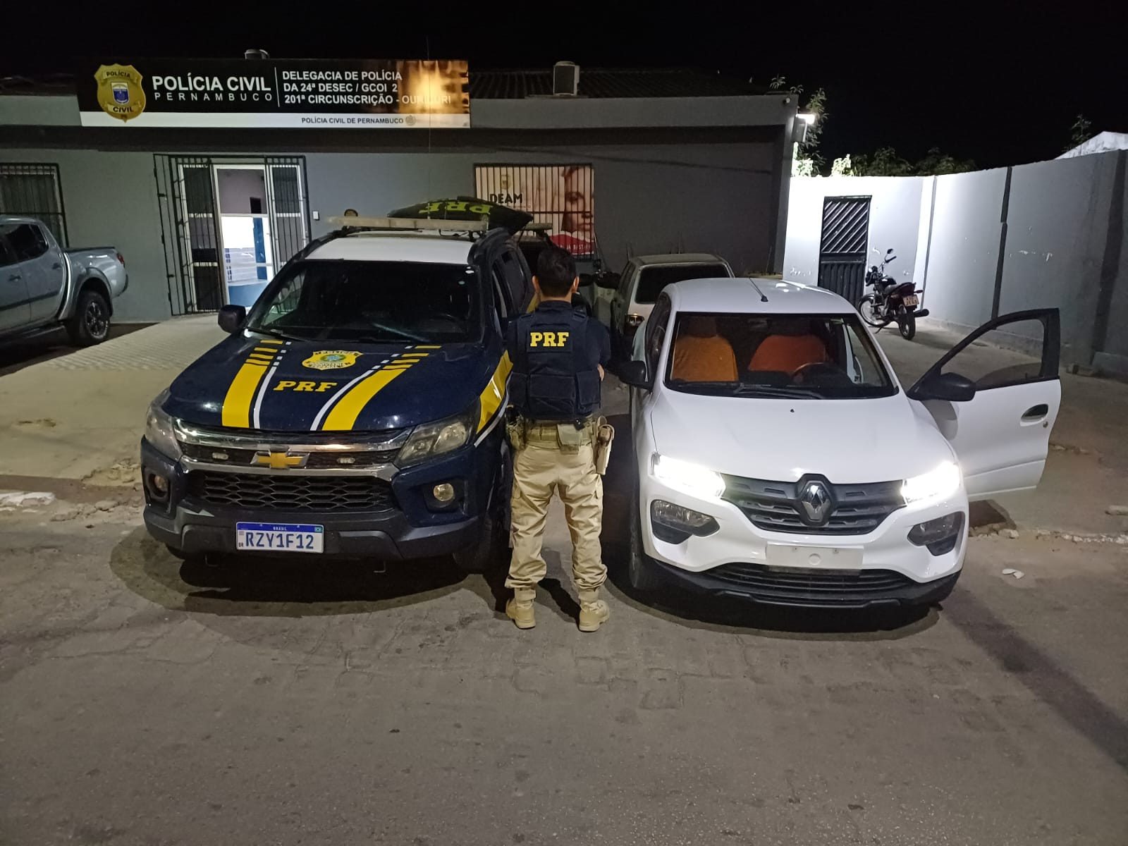 Fotografia noturna em frente a uma delegacia da Polícia Civil de Pernambuco. No centro da imagem, um agente da Polícia Rodoviária Federal (PRF), de costas, veste uniforme tático azul-marinho, com a sigla PRF em amarelo nas costas, calça bege e botas. Ele está entre duas viaturas: à esquerda, uma caminhonete azul-escura da PRF, com faixas amarelas e identificação oficial; à direita, um carro branco da Renault, com os faróis acesos e a porta dianteira do passageiro aberta. Ao fundo, a fachada da delegacia, com a identificação da Polícia Civil de Pernambuco, além de outros veículos estacionados. A cena é iluminada artificialmente durante a noite.
