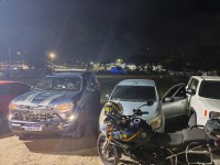Foto noturna mostrando uma viatura e uma motocicleta da PRF ao lado de um carro prata com a porta aberta. Os veículos estão estacionados em uma área iluminada, com árvores e luzes da cidade ao fundo. A cena sugere uma fiscalização policial.