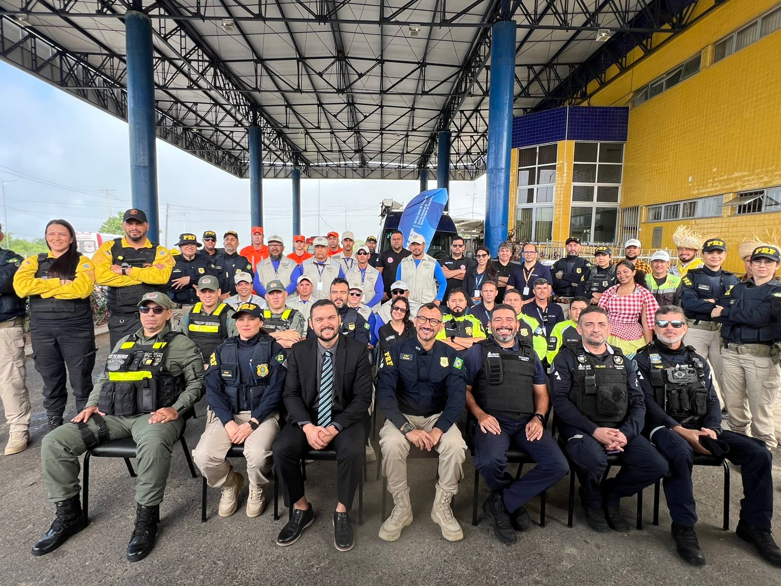 Foto de grupo com cerca de 50 pessoas, entre agentes de segurança e outros profissionais, reunidas em frente a um prédio com colunas azuis e fachada amarela. Alguns estão sentados na primeira fila e outros em pé atrás. A maioria sorri para a câmera, em um registro de caráter institucional ou operacional.