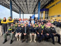 Foto de grupo com cerca de 50 pessoas, entre agentes de segurança e outros profissionais, reunidas em frente a um prédio com colunas azuis e fachada amarela. Alguns estão sentados na primeira fila e outros em pé atrás. A maioria sorri para a câmera, em um registro de caráter institucional ou operacional.