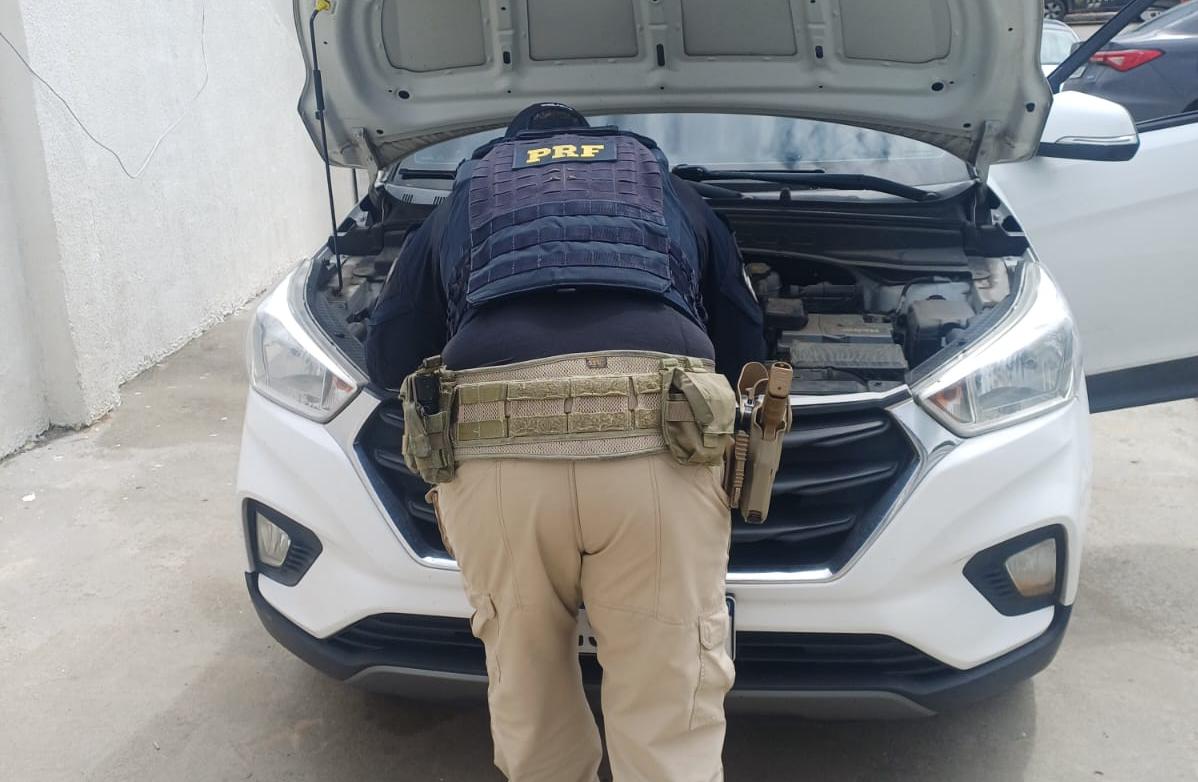 Policial Rodoviário Federal realiza inspeção no compartimento do motor de um veículo SUV branco com o capô aberto. O agente está de costas para a câmera, utilizando uniforme da PRF e equipamento tático. A imagem foi registrada em uma área externa pavimentada, próxima a um muro claro, durante uma fiscalização veicular.