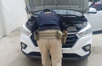 Policial Rodoviário Federal realiza inspeção no compartimento do motor de um veículo SUV branco com o capô aberto. O agente está de costas para a câmera, utilizando uniforme da PRF e equipamento tático. A imagem foi registrada em uma área externa pavimentada, próxima a um muro claro, durante uma fiscalização veicular.
