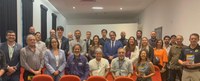 Fotografia de grupo realizada em um auditório durante a reunião do Fórum de Gestores Federais, no Recife. Cerca de 30 pessoas aparecem posando para a foto, distribuídas em três fileiras entre cadeiras vermelhas. Os participantes incluem homens e mulheres de diferentes idades, vestindo roupas sociais e trajes institucionais. Ao centro, estão representantes de órgãos federais e do sistema de segurança pública. Alguns seguram exemplares de uma publicação. O ambiente possui paredes claras, cortinas escuras nas janelas e iluminação interna. Os participantes estão voltados para a câmera, em clima formal e colaborativo, simbolizando a integração entre instituições federais que atuam em Pernambuco.