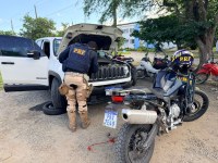 Fotografia registrada em uma área externa durante uma fiscalização da Polícia Rodoviária Federal (PRF). Em primeiro plano, um policial rodoviário federal, visto de costas e usando colete com a identificação “PRF”, realiza uma inspeção no compartimento do motor de um veículo utilitário branco com o capô aberto. Ao lado do veículo, há um pneu apoiado no chão. Em destaque à direita, uma motocicleta operacional da PRF está estacionada, com capacete institucional sobre o banco. Ao fundo, aparecem outras motocicletas estacionadas, árvores de grande porte e uma edificação de paredes brancas e detalhes em azul. O ambiente é iluminado pela luz do dia e transmite uma ação de fiscalização e verificação veicular.