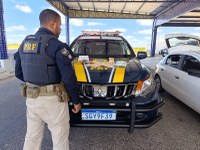 Um policial rodoviário federal, visto de costas, está ao lado de uma viatura da PRF em uma área de fiscalização coberta. Sobre o capô da viatura há dinheiro e outros materiais apreendidos expostos. À direita, um carro branco está estacionado com o porta-malas aberto. A cena registra uma ação de fiscalização da PRF durante o dia.