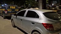 Foto noturna mostrando um carro prata estacionado à frente de uma viatura da PRF. Os veículos estão parados em uma área urbana iluminada, com árvores e outros carros ao fundo. A imagem registra uma abordagem ou fiscalização realizada pela Polícia Rodoviária Federal.