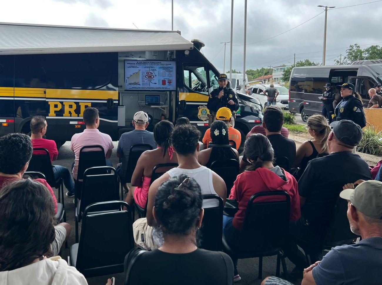 Pessoas sentadas assistem a uma palestra da PRF realizada ao lado de uma unidade móvel da corporação