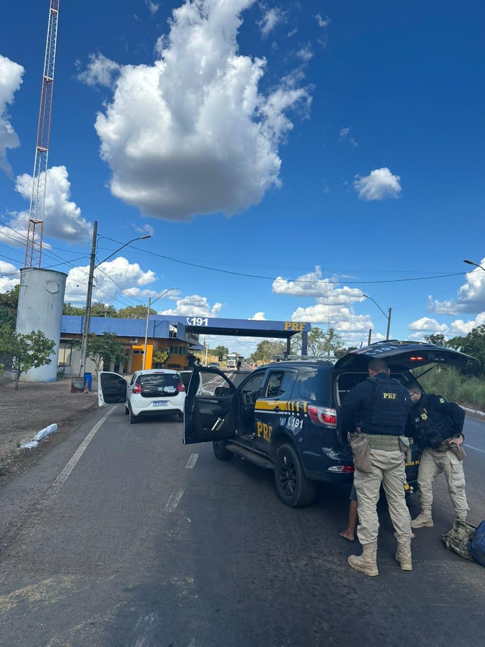 A imagem mostra uma ação de fiscalização da Polícia Rodoviária Federal (PRF) em uma rodovia. Em primeiro plano, há uma viatura caracterizada da PRF estacionada na pista, com o porta-malas e as portas abertas. Dois policiais realizam uma abordagem e revista em uma pessoa posicionada ao lado da viatura. À frente da viatura, aparece um veículo de passeio branco também parado no acostamento, com a porta do motorista aberta. Ao fundo, é possível ver a estrutura de uma unidade operacional da PRF, identificada pelo número 191 e pela sigla PRF sobre um pórtico que atravessa a rodovia. O cenário é diurno, com céu azul e diversas nuvens brancas de grande porte, proporcionando boa visibilidade. Há postes de iluminação, fiação aérea e vegetação nas margens da estrada. A imagem transmite a ideia de uma operação de fiscalização e segurança viária realizada pela PRF.