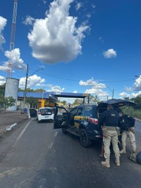 A imagem mostra uma ação de fiscalização da Polícia Rodoviária Federal (PRF) em uma rodovia. Em primeiro plano, há uma viatura caracterizada da PRF estacionada na pista, com o porta-malas e as portas abertas. Dois policiais realizam uma abordagem e revista em uma pessoa posicionada ao lado da viatura.  À frente da viatura, aparece um veículo de passeio branco também parado no acostamento, com a porta do motorista aberta. Ao fundo, é possível ver a estrutura de uma unidade operacional da PRF, identificada pelo número 191 e pela sigla PRF sobre um pórtico que atravessa a rodovia.  O cenário é diurno, com céu azul e diversas nuvens brancas de grande porte, proporcionando boa visibilidade. Há postes de iluminação, fiação aérea e vegetação nas margens da estrada. A imagem transmite a ideia de uma operação de fiscalização e segurança viária realizada pela PRF.