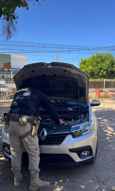 Descrição da imagem: Policiais rodoviários federais realizam inspeção no compartimento do motor de um Renault prata, com o capô aberto, durante fiscalização em via urbana. Ao fundo, há um prédio público, árvores e céu limpo.
