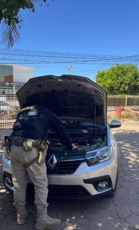 Descrição da imagem: Policiais rodoviários federais realizam inspeção no compartimento do motor de um Renault prata, com o capô aberto, durante fiscalização em via urbana. Ao fundo, há um prédio público, árvores e céu limpo.