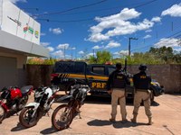 **Descrição acessível:** Dois policiais rodoviários federais observam motocicletas apreendidas ao lado de uma viatura da PRF, em frente a um prédio público. A cena ocorre durante o dia, sob céu azul com nuvens, em ambiente que sugere uma ação de fiscalização ou operação policial..