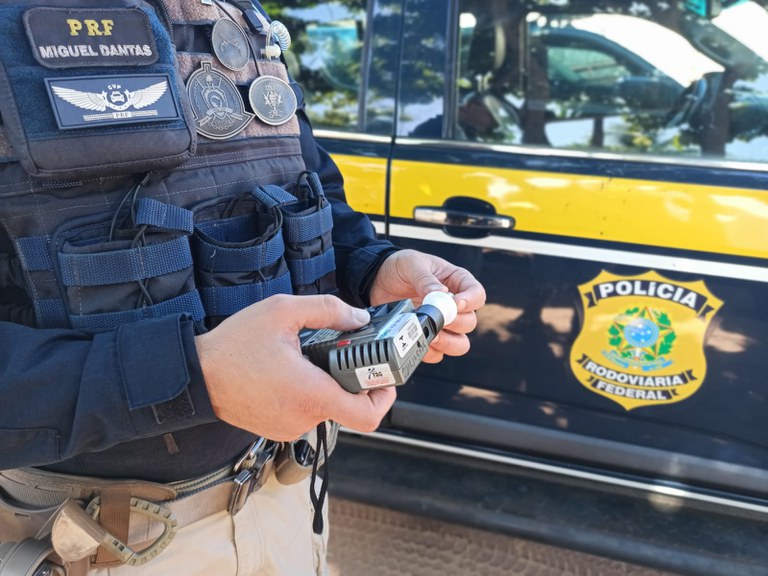 PRF segurando um etilometro e ao fundo uma viatura caracterizada