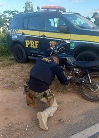 Um PRF examinando a motocicleta adulterada. Ao fundo uma viatura.