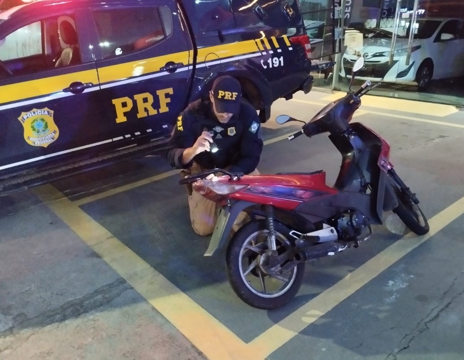 Motocicleta recuperada com o policial inspecionando em frente a viatura da PRF