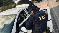 Policial rodoviário federal, de costas e fardado, vistoria o interior de um carro com a porta aberta na rodovia.