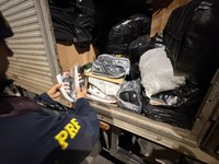 Policial Rodoviário Federal examina um par de tênis durante fiscalização em um caminhão de carga. No interior do compartimento, são observadas caixas de calçados, pares de tênis embalados individualmente e diversos volumes acondicionados em sacos pretos. A imagem foi registrada durante a noite e evidencia a conferência de mercadorias transportadas.