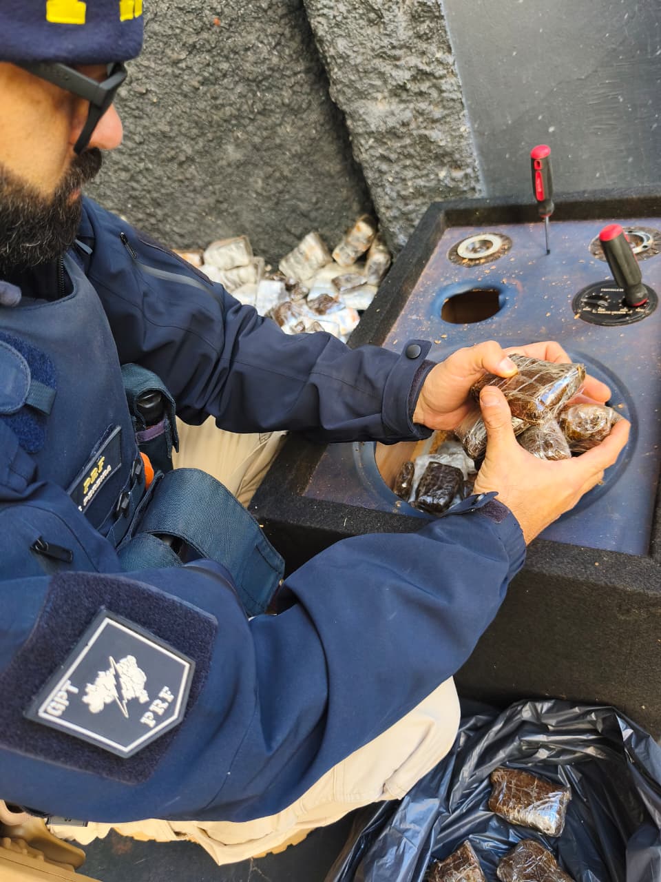 Policial rodoviário federal remove volumes de haxixe encontrados em compartimentos ocultos durante inspeção veicular.