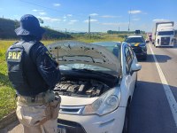 A imagem mostra uma fiscalização da Polícia Rodoviária Federal (PRF) realizada durante o dia em uma rodovia. Em primeiro plano, um policial rodoviário federal, visto de costas e uniformizado, está ao lado de um automóvel branco parado no acostamento. O capô do veículo está aberto, expondo o compartimento do motor para inspeção.
