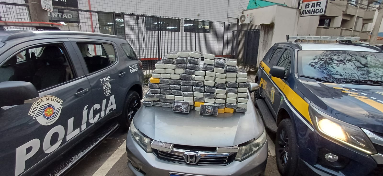 Grande quantidade de tabletes de droga empilhados sobre o capô de um carro prata, posicionados entre uma viatura da Polícia Militar e uma viatura da PRF. A imagem foi registrada em área urbana durante o dia.