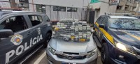 Grande quantidade de tabletes de droga empilhados sobre o capô de um carro prata, posicionados entre uma viatura da Polícia Militar e uma viatura da PRF. A imagem foi registrada em área urbana durante o dia.