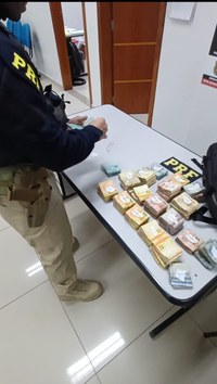 Policial rodoviário federal realiza a contagem de dinheiro sobre uma mesa com diversas pilhas de cédulas organizadas. A cena ocorre no interior de uma unidade da PRF, com portas e mobiliário de escritório ao fundo