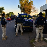 Imagem institucional da Polícia Rodoviária Federal (PRF) mostrando três policiais rodoviários federais posicionados ao redor de uma viatura com luzes de emergência azuis acionadas. A cena ocorre em uma via urbana durante o entardecer, com veículos trafegando ao fundo e árvores iluminadas pela luz do pôr do sol.
