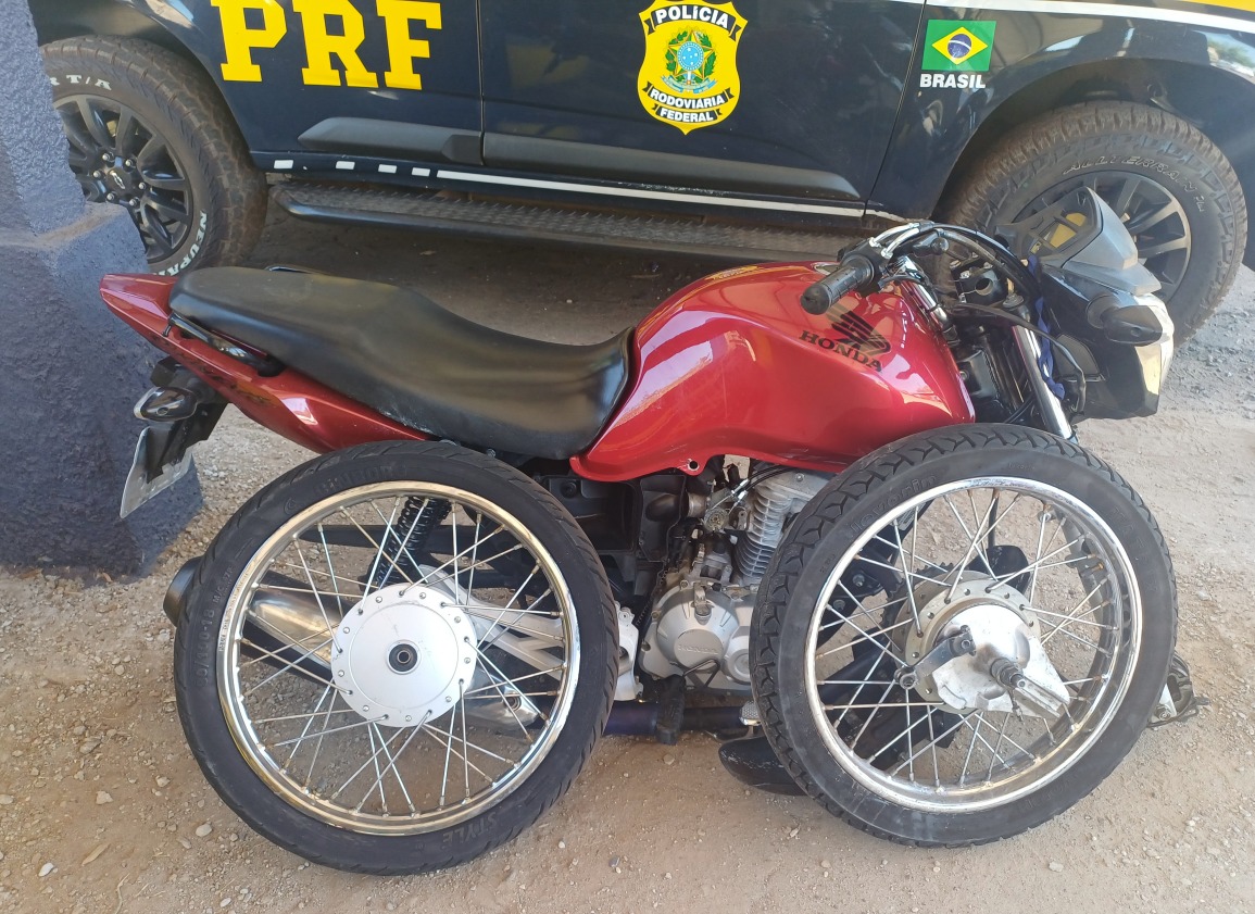 Imagem institucional da Polícia Rodoviária Federal (PRF) mostrando uma motocicleta vermelha posicionada em frente a uma viatura da PRF. A motocicleta aparece sem a roda dianteira instalada e com duas rodas removidas apoiadas ao lado do veículo. Ao fundo, é possível visualizar parcialmente a lateral da viatura oficial da PRF.