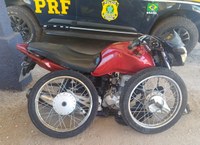Imagem institucional da Polícia Rodoviária Federal (PRF) mostrando uma motocicleta vermelha posicionada em frente a uma viatura da PRF. A motocicleta aparece sem a roda dianteira instalada e com duas rodas removidas apoiadas ao lado do veículo. Ao fundo, é possível visualizar parcialmente a lateral da viatura oficial da PRF.